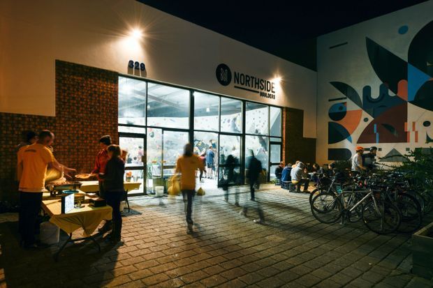 Night-time exterior view of a contemporary commercial bouldering gym entrance. The illuminated glass storefront displays the Northside Boulders sign, with people gathered around outdoor tables, bicycles parked beside the facade, brick paving, and a large colourful geometric mural on the adjoining wall.