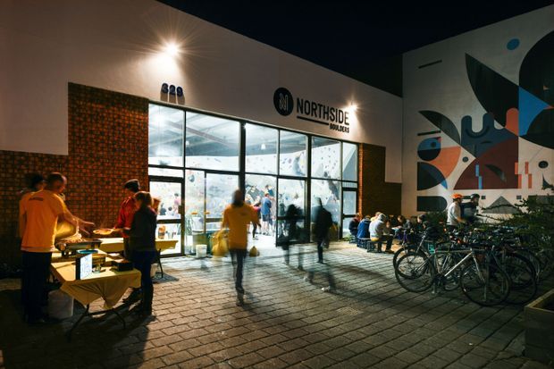 Night-time exterior view of the Northside Boulders climbing gym, with a brightly illuminated glazed entrance, signage, outdoor tables and people, rows of bicycles, and a large colourful geometric mural on the facade. The building is set around a paved public forecourt.