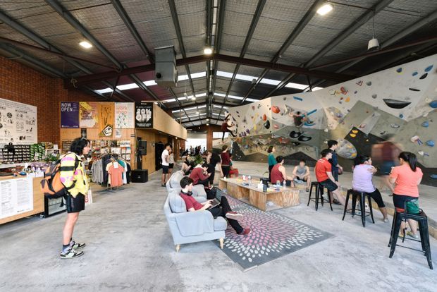 A spacious indoor commercial recreation venue with an industrial warehouse character. The open-plan space has exposed steel roof framing, corrugated metal roofing, concrete floors, exposed brickwork, café-style seating, a retail counter and clothing displays, and a large indoor climbing wall with colorful climbing holds. Visitors gather around tables and lounge seating beneath bright overhead lighting.