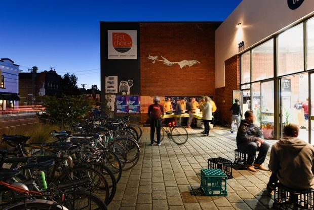 Dusk exterior view of a contemporary urban commercial venue, with a brick and dark-panel facade, large glazed shopfront windows, a paved public forecourt, outdoor seating, bicycles parked along the street, and people gathered near the entrance. Illuminated signage and interior activity create a lively streetscape atmosphere.