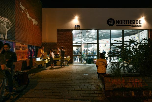 Night-time exterior view of a contemporary brick commercial building entrance branded “Northside Boulders.” The forecourt is paved with brick, with large glazed windows revealing an active climbing gym interior, while people, bicycles, market tables and planted raised beds occupy the entrance area. Warm exterior lighting contrasts with the illuminated interior.
