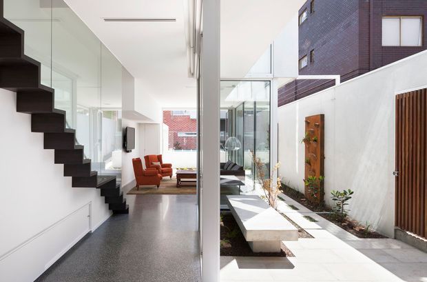 Bright contemporary multi-level residential interior viewed toward an open-plan living area and glazed courtyard. A dramatic dark floating staircase runs along the left wall, while full-height glazing connects the living space to a narrow landscaped courtyard with white paving, raised concrete seating or planters, and a timber slatted gate. The interior has white walls, polished grey flooring, extensive glass, and warm red lounge chairs.