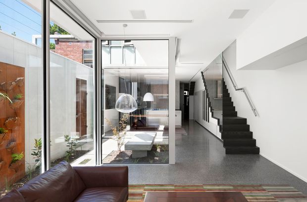 A bright contemporary residential interior showing an open-plan living area and hallway with a dark leather sofa in the foreground, a glazed internal courtyard or lightwell, polished grey flooring, and a sculptural black staircase along a white wall. Floor-to-ceiling glazing brings in abundant natural light, while exposed brick and timber elements are visible around the courtyard.