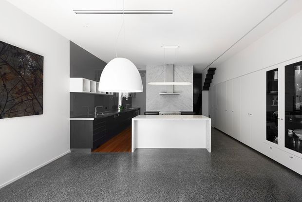 A bright, minimalist open-plan kitchen viewed from the living area. The space features a white island with a dark benchtop, black cabinetry along the rear wall, a large white dome pendant, integrated white storage cabinetry with glass-front display sections, polished grey flooring, and a stairway visible at the rear.