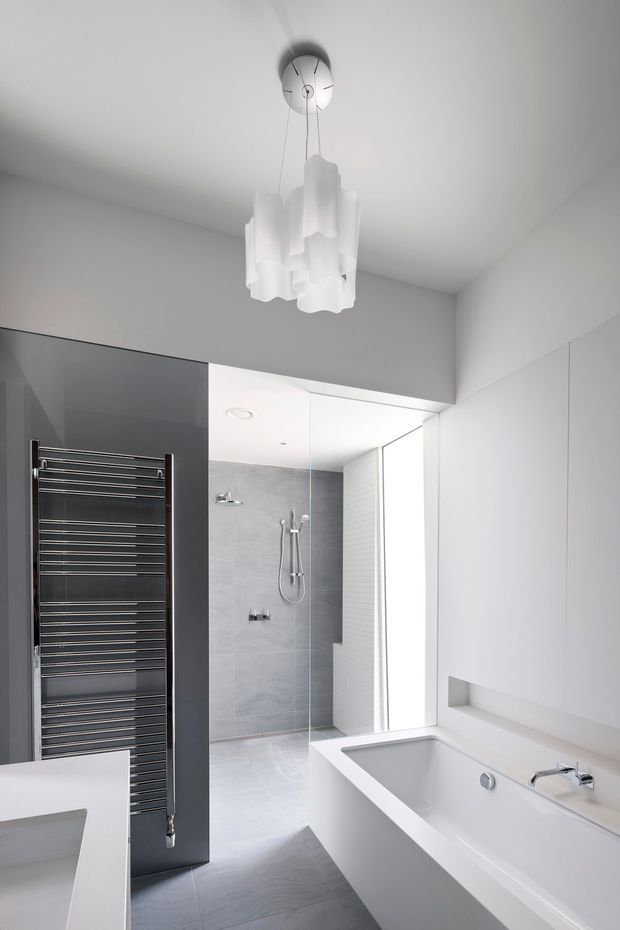 Bright, minimalist contemporary bathroom with a freestanding white bathtub, wall-mounted chrome tapware, a walk-in glass shower, pale grey tiled shower walls and floor, a dark charcoal heated towel rail feature panel, and white built-in cabinetry with an open recessed shelf. A sculptural white pendant light hangs from the ceiling, adding a soft decorative element.