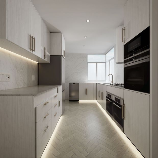 A bright, narrow contemporary galley kitchen with extensive pale vertical-grain cabinetry, integrated black appliances, marble-look wall and benchtop surfaces, under-cabinet lighting, and a light herringbone floor. Full-height glazing at the far end brings in natural light, while concealed LED lighting outlines the cabinetry and floor edges.