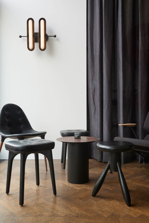 A refined interior vignette featuring black leather lounge chairs and stools arranged around a small round dark side table. The room has a white wall with a sculptural wall sconce, dark full-height curtains, and a warm parquet timber floor. The overall composition is understated, moody, and contemporary with strong mid-century-inspired furniture forms.