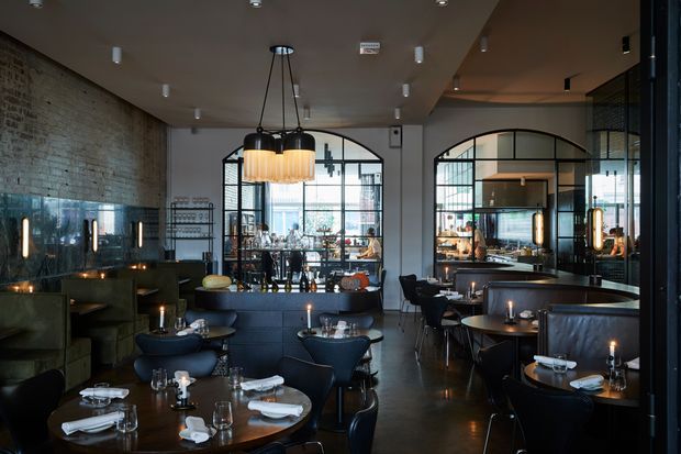 A dimly lit contemporary restaurant interior with dark dining tables and chairs, banquette seating, exposed brick walls, black-framed industrial-style arched glazed partitions, and a large cluster pendant light over the central seating area. The space includes a visible bar or service area beyond the partitions, reflective dark flooring, and warm ambient lighting.
