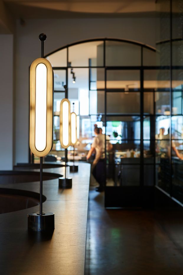 A moody commercial interior, likely a café, bar, or restaurant, featuring a long dark counter with sculptural vertical pendant lamps. Black-framed glass partitions with curved upper sections divide the space, while warm ambient lighting, polished dark flooring, and blurred patrons create a refined contemporary atmosphere.