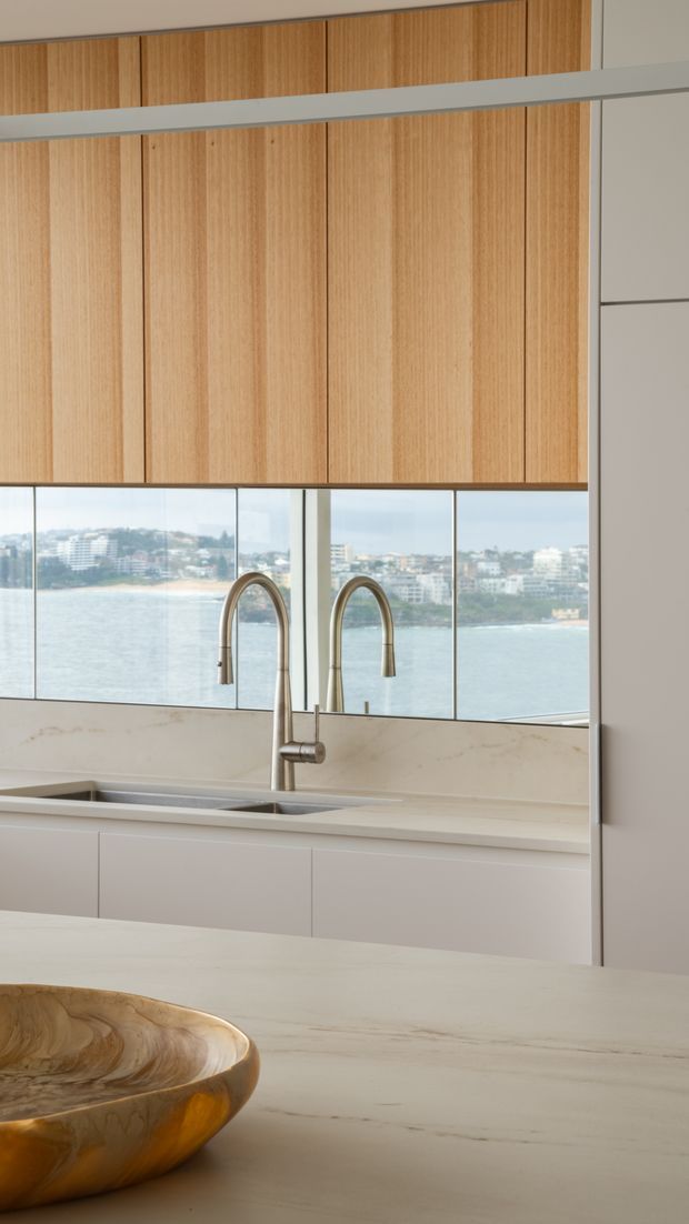 A refined contemporary kitchen overlooking the water. The image focuses on a pale stone island with a sculptural wooden bowl, a minimalist integrated sink and twin brushed-metal gooseneck taps, light neutral cabinetry, and full-height glazing. Vertical natural timber overhead cabinetry adds warmth, while the coastal view is visible beyond the splashback glazing.