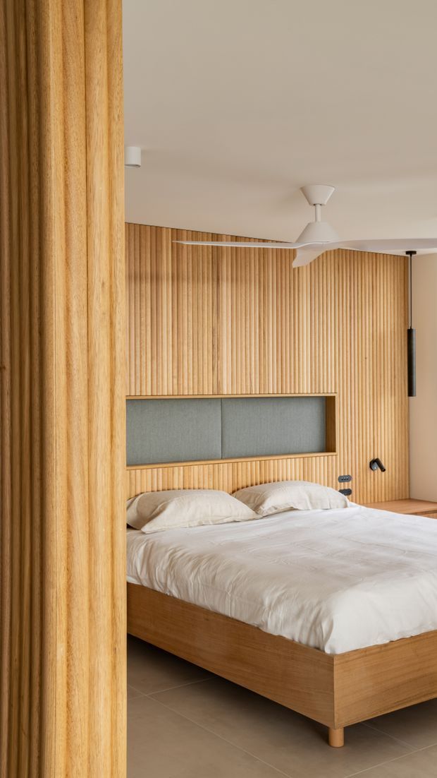 A warm, contemporary bedroom viewed through a timber-lined opening. The room features a low platform bed with white bedding, an integrated grey upholstered headboard framed in timber, extensive vertical timber wall panelling, a neutral ceiling, tiled flooring, a white ceiling fan, and small wall-mounted reading lights.