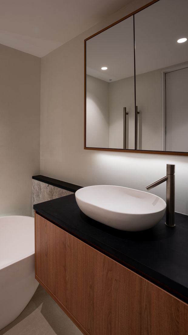 A compact contemporary bathroom featuring a wall-mounted timber vanity with flat-panel cabinetry, a dark stone-look benchtop, white vessel basin, and brushed metal tapware. A large framed mirrored cabinet spans the wall above the vanity, with warm recessed lighting and neutral beige wall and floor finishes. The edge of a freestanding or built-in white bathtub is visible at left.