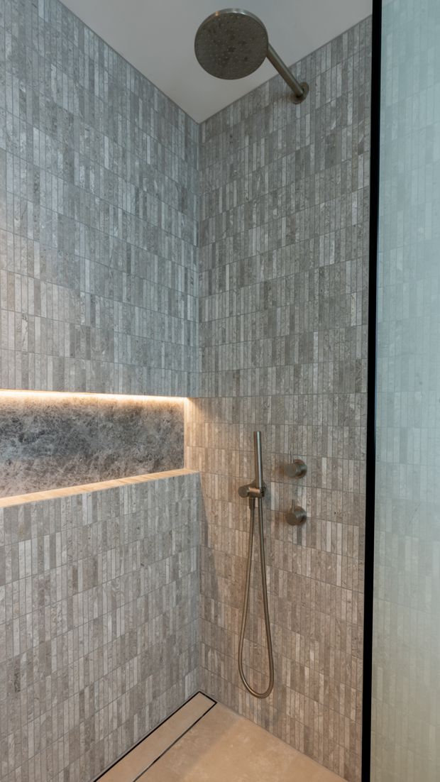 A modern walk-in shower with full-height vertically patterned grey stone-look tiles, a large round overhead shower, handheld shower, and recessed built-in bench niche with warm LED lighting. A frameless glass screen with a dark edge is visible at the right. The space has a refined, spa-like appearance with neutral grey and beige tones.