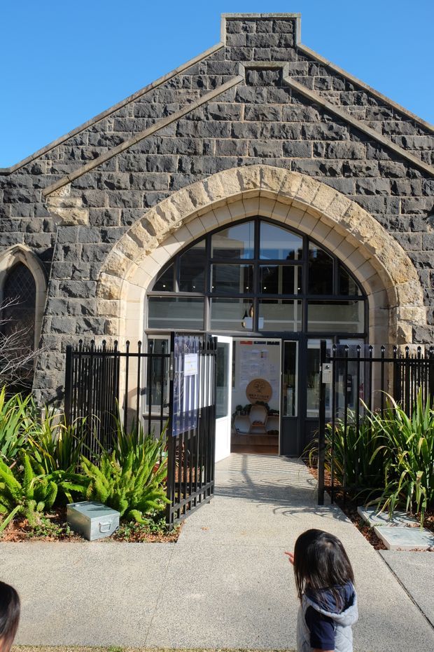 Exterior view of a historic stone building entrance with a steep gabled facade, dark rough-cut masonry, contrasting pale stone arched surround, and large black-framed glazed entry doors. A black metal fence and landscaped planting line the accessible paved approach.