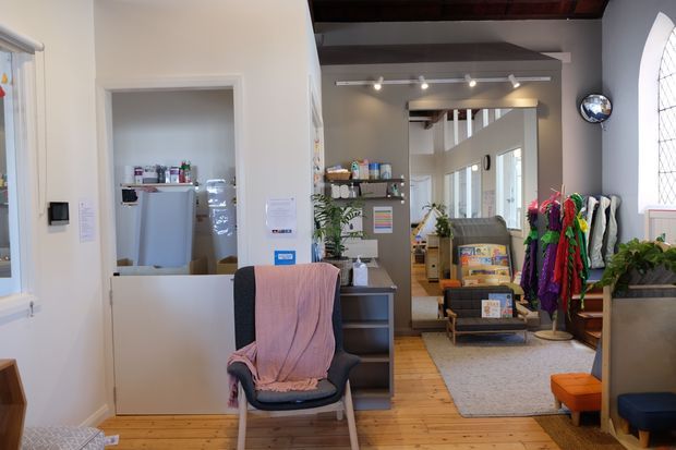 A bright, eclectic commercial interior, likely a reception or waiting area. The foreground contains a chair, service counter, and open pass-through to a back workspace or kitchenette. Exposed dark timber ceiling beams, timber floorboards, grey painted walls, track lighting, shelving, plants, colourful garments or textiles, and a large arched leaded-glass window define the space.