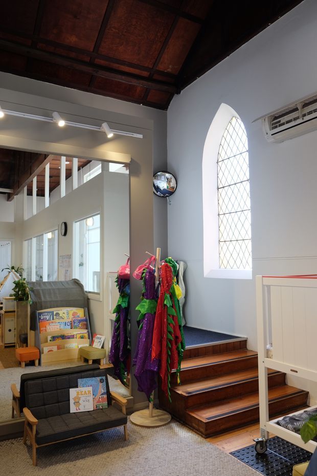 A bright, compact interior reading and activity area with a small lounge chair, children's book display, colourful dress-up clothing rack, carpeted floor, and a short flight of timber stairs leading to a raised level. White and grey walls feature a tall arched leaded-glass window, while the exposed dark timber ceiling and beams add strong architectural character. A partial mezzanine or upper-level opening is visible beyond the stairs.