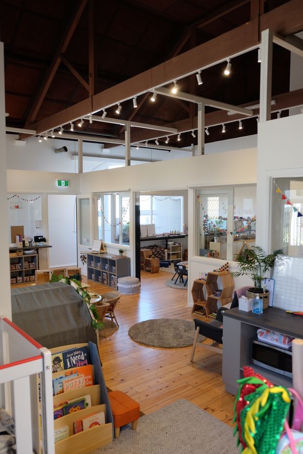 Interior view of a bright, open-plan commercial or community space, likely a childcare or educational facility. The room has exposed dark timber roof framing and a high ceiling, white partition walls with large internal windows, warm timber floorboards, extensive track lighting, open shelving, books, children's furniture and play materials. The space feels welcoming and functional, with several interconnected activity areas visible through glazed partitions.
