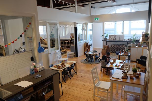 Bright, open-plan community or educational interior with timber flooring, multiple small dining and activity tables, shelving, a kitchenette or service counter, and large windows. The space has a casual, welcoming layout with playful bunting, potted plants, and track lighting.