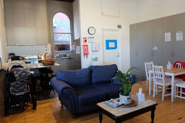 A bright, open-plan communal interior combining a compact kitchen, lounge, and dining area. The room has high ceilings, warm timber floorboards, pale tiled kitchen walls, extensive grey built-in cabinetry, a navy upholstered sofa, and a traditional timber coffee table. A dining table with white chairs sits beside the cabinetry, while a large arched window brings in natural light. The space appears functional and institutional or shared rather than a private residential room.