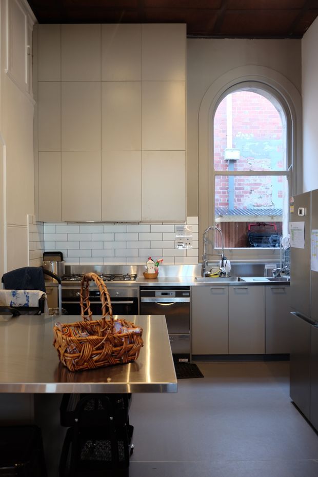 A compact, functional kitchen with a stainless-steel island and integrated appliances, matte grey lower cabinetry, tall cream upper cabinetry, and a white subway-tile splashback. A large arched window provides natural light, while a dark timber-lined ceiling adds warmth. A woven basket sits on the island.