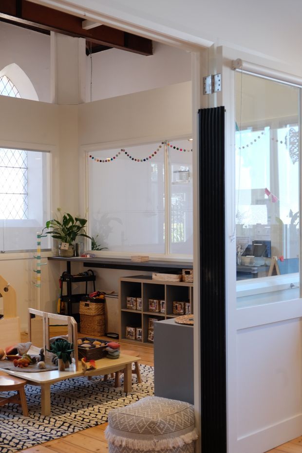 A bright, eclectic interior viewed through a partially open glazed door, showing a casual family living or play area with timber flooring, a patterned rug, low wooden tables, children’s toys, baskets, shelving, and indoor plants. The room has white walls, large internal windows, decorative bunting, a black vertical radiator, and visible exposed dark timber framing near the ceiling. A second workspace or room is visible through the glazing on the right.