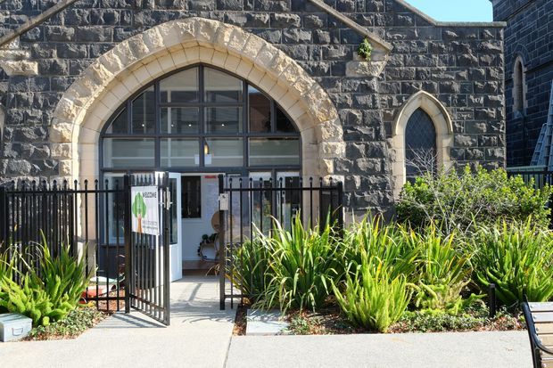 Exterior view of a dark stone masonry building with prominent pale stone pointed-arch surrounds, a large arched glazed entrance, a smaller pointed-arch window, black metal fencing and gate, and landscaped plantings along the concrete approach.