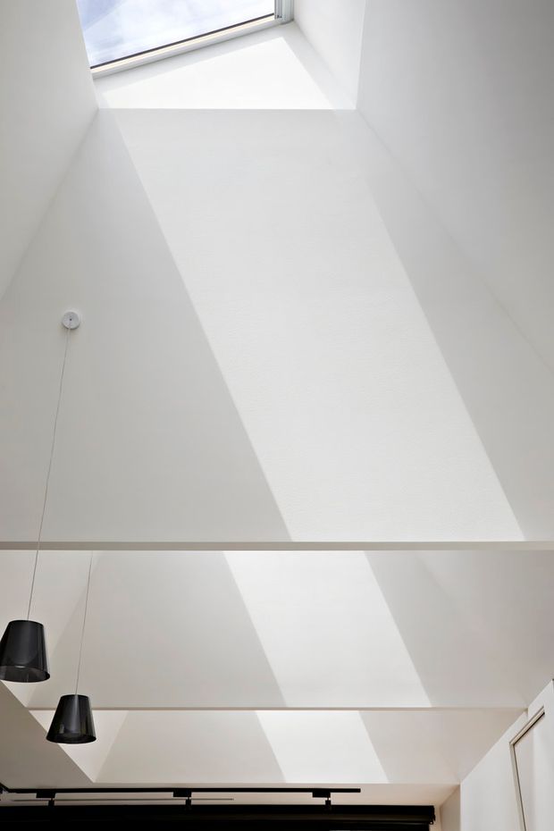 Upward-looking view of a bright white interior ceiling with a large roof skylight or clerestory opening casting strong geometric shafts of daylight across the sloped ceiling. Two black pendant lights hang at the left, with slim horizontal structural or decorative beams spanning the space and dark window covering visible along the lower edge. The scene is minimalist and architectural, focused on the ceiling, natural light, and roof glazing.