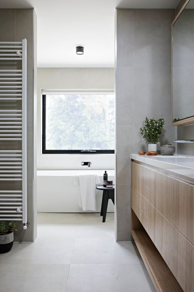 A bright, contemporary bathroom with a freestanding white bathtub positioned beneath a large window. The space features pale grey large-format tiled walls and flooring, a timber-look floating vanity with a white benchtop, open shelving, a black-framed mirror cabinet, and a slim white heated towel rail. Minimal black fixtures and a small plant add contrast.