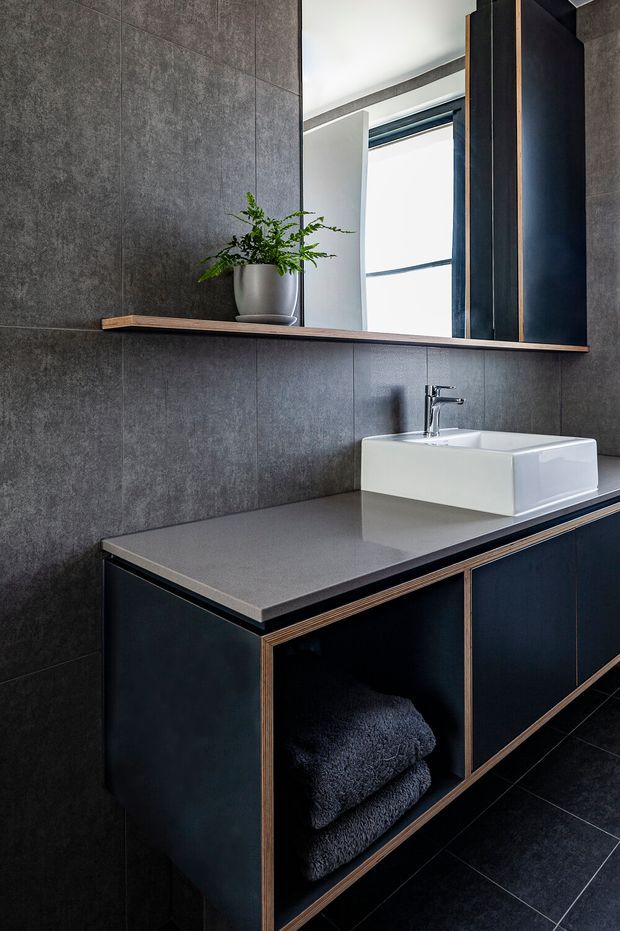 A refined modern bathroom vanity viewed straight on. The room features dark charcoal textured wall and floor tiles, a dark blue-black floating vanity with warm timber edging, a smooth grey benchtop, and a white vessel basin with chrome tapware. A large framed mirror spans the vanity, reflecting a bright window and a potted plant. Open shelving beneath the vanity holds folded dark towels.