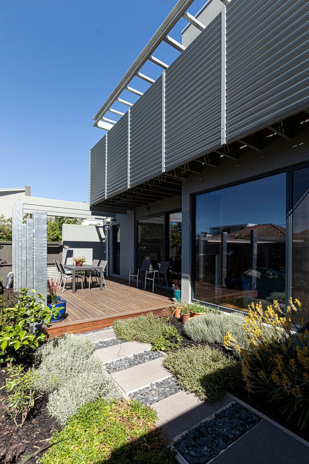 Exterior view of a contemporary residence with a timber deck, outdoor dining area, large dark-framed glazed doors, and prominent horizontal metal louvre sunshades beneath a pergola-like roof structure. A landscaped garden with low silvery shrubs, gravel strips, stepping slabs, and potted plants leads toward the deck. Bright clear daylight emphasizes the crisp modern facade and strong shadow lines.
