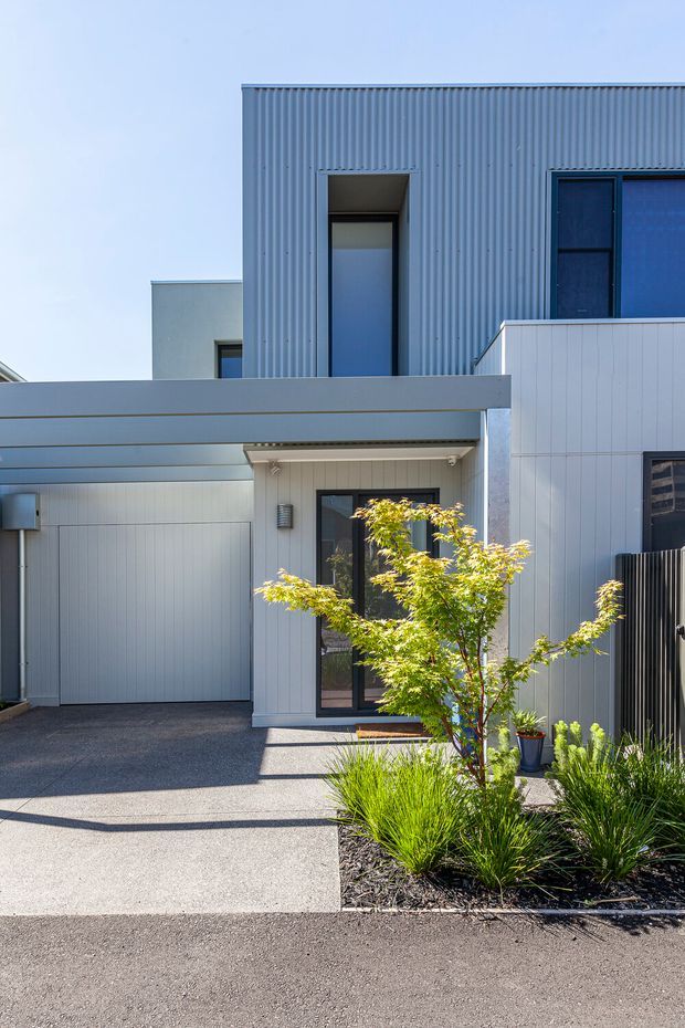 Exterior view of a contemporary two-storey home with a corrugated blue-grey upper facade, pale vertical-panel cladding, dark-framed windows and doors, and an attached covered carport. A paved driveway and landscaped garden bed with ornamental grasses and a small tree occupy the foreground.