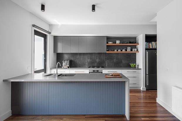 Contemporary kitchen with a large island featuring vertically fluted blue-grey cabinetry and a light stone benchtop. The room has dark grey upper cabinets, open timber shelves, a dark stone-look splashback, integrated appliances, warm timber flooring, white walls and ceiling, black-framed glazing, and recessed black cylindrical ceiling lights.