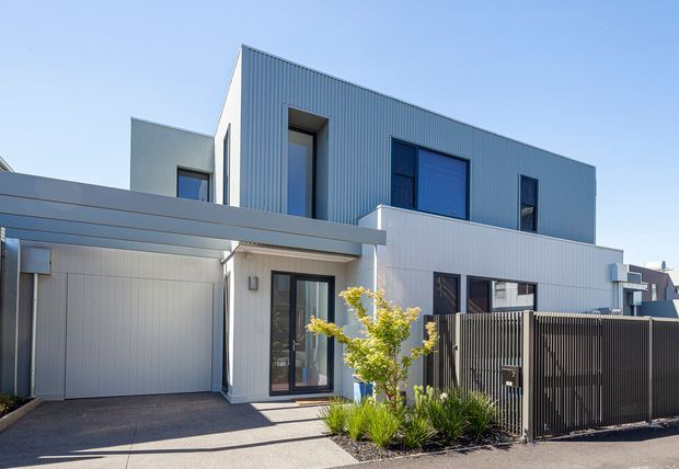 A contemporary two-storey residential facade with clean rectilinear volumes, white and grey vertically ribbed metal cladding, dark-framed windows, and a projecting flat-roofed carport or garage structure. The frontage includes a large garage door, entry door, dark vertical-slat privacy fencing, paved driveway, and a small landscaped garden bed. Bright daylight highlights the crisp modern exterior.