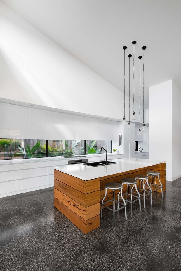 Bright, spacious contemporary kitchen with a large white island featuring a timber-clad base, integrated sink and black tap, extensive white cabinetry, a long horizontal window, and multiple sculptural pendant lights suspended from a high ceiling. The dark polished speckled floor contrasts with the predominantly white interior, while greenery is visible outside the glazing.