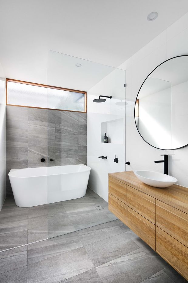 Bright contemporary bathroom featuring a freestanding white bathtub, large-format grey stone-look wall and floor tiles, frameless glass shower enclosure, black fixtures, a round black-framed mirror, and a floating light timber vanity with a white vessel basin. A high frosted window brings in natural light.