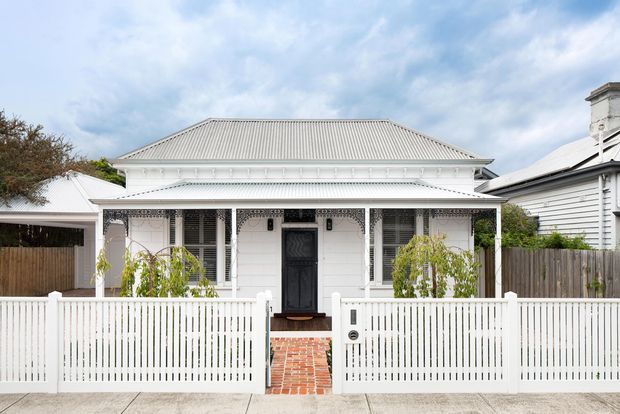 Front exterior view of a compact, single-storey heritage-style residence with a symmetrical white facade, corrugated hipped roof, decorative verandah detailing, dark central entry door, and a white picket fence. A brick pathway leads from the footpath to the entrance, with neighbouring residential structures visible at either side.