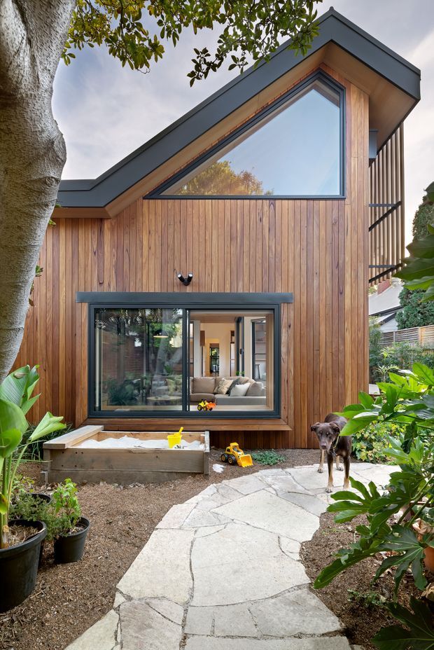 Exterior view of a compact contemporary timber-clad extension with a sharply angled roofline, large triangular window, dark metal trims, and lower glazed doors/windows. A pale irregular stone pathway leads through a landscaped garden to the building, with a small raised sandpit and children's toys in the foreground. The interior living area is visible through the glazing.