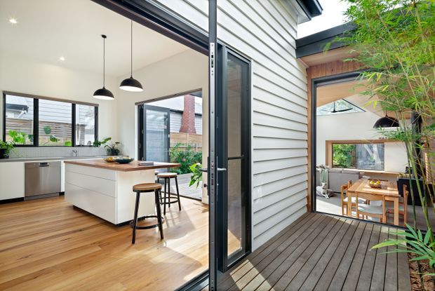 Bright contemporary open-plan kitchen and dining area connected by large glazed doors to a timber deck and planted courtyard. The kitchen features white flat-panel cabinetry, a timber island benchtop, dark-framed windows and doors, pendant lighting, and timber flooring. A dining space is visible beyond the deck, with indoor-outdoor connectivity and a planted bamboo screen.
