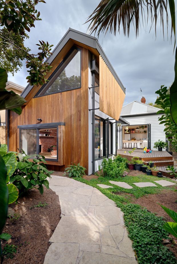 Exterior view of a contemporary timber-clad extension or small pavilion in a leafy courtyard garden. The building has a steeply pitched gable form, a dramatic triangular window, vertical natural timber cladding, dark metal window and door frames, and a raised timber deck connecting to an older light-coloured weatherboard house. A paved path and landscaped planting lead toward the structure.