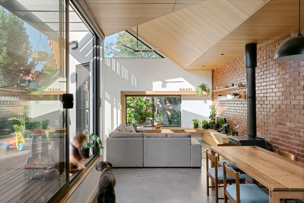 Bright open-plan living and dining space with a contemporary extension feel, featuring full-height glazing, a warm timber-lined angled ceiling, exposed red brick wall, built-in timber seating and cabinetry, a grey sectional sofa, and a black freestanding wood-burning stove. A dining table with stools occupies the foreground, while the living area opens toward a garden view.