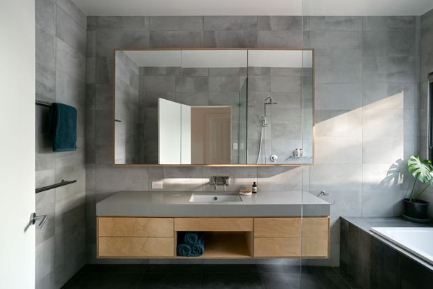 Contemporary bathroom with large-format grey tiled walls and floor, a floating timber vanity with integrated basin and grey benchtop, mirrored wall cabinetry, frameless glass shower enclosure, and a bathtub at right. Natural light and a potted plant add a calm, refined feel.