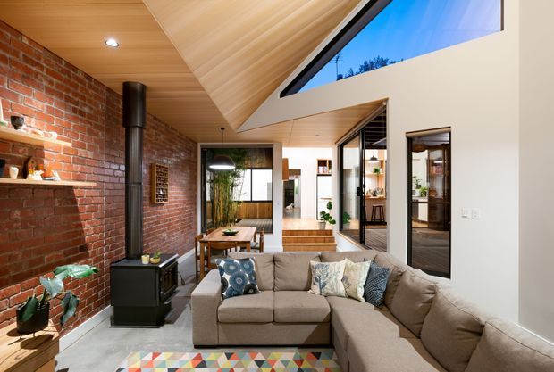 A warm, contemporary living room with a large L-shaped beige sectional sofa, exposed red brick feature wall, black wood-burning stove, timber-lined pitched ceiling, and clerestory glazing. The room opens toward a raised dining or circulation area, with indoor plants and colorful patterned cushions and rug adding contrast.