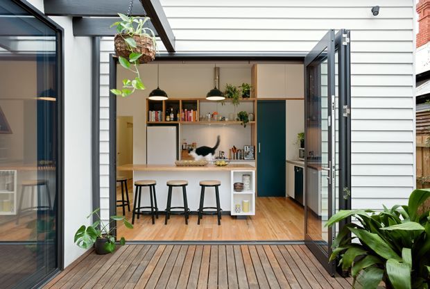 View from a timber deck into a bright, compact kitchen and dining space through wide-open glazed doors. The room features white horizontal weatherboard walls, warm timber flooring, a white island with four black stools, open timber shelving, pendant lights, indoor plants, and a deep blue door. A glazed adjoining space is visible to the left, while exterior greenery frames the deck.