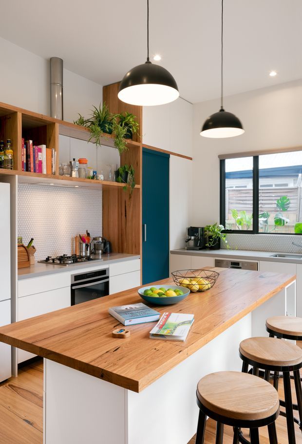 Bright, contemporary kitchen featuring a large timber island with white cabinetry and timber stools, open timber shelving, white cabinetry, a white hexagonal tile splashback, stainless-steel appliances, teal cabinetry, indoor plants, and two black pendant lights. A window provides natural light over the rear benchtop.