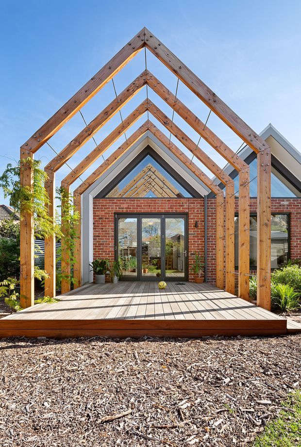 Exterior view of a contemporary residential extension or pavilion with a dramatic series of exposed timber triangular frames forming a covered entry and deck. The structure combines red brick walls, dark-framed glazed double doors, grey metal roof elements, and landscaped planting around a timber deck. Bright daylight and strong geometric repetition create a bold architectural focal point.