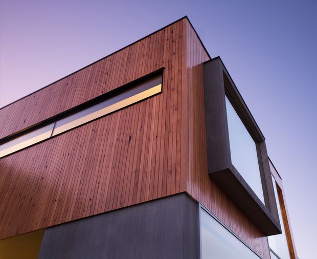 Low-angle exterior detail of a contemporary multi-storey building with vertical timber cladding, dark projecting window surrounds and large glazed openings. The sharply folded roofline and clean geometric forms are silhouetted against a purple evening sky.