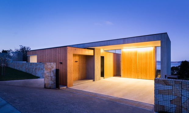 Contemporary single-storey residential facade at dusk, featuring a long low-slope roofline, warm timber-clad recessed entry and garage doors, exposed concrete framing, a broad illuminated driveway or carport-like opening, and gabion-style stone retaining walls. The composition emphasizes the building exterior rather than an interior room.