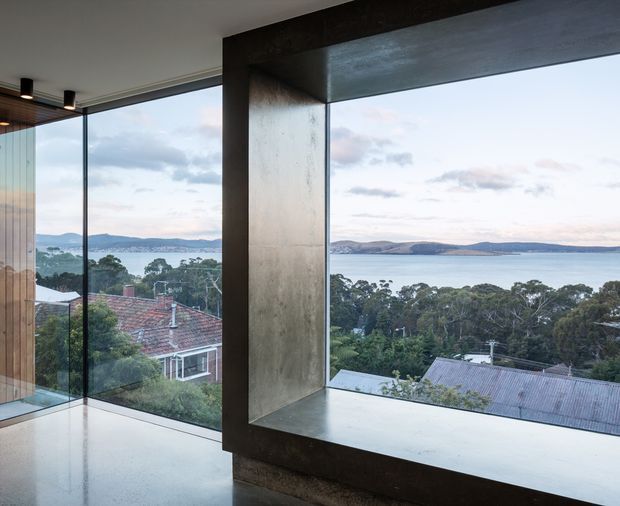 Interior architectural view dominated by a large corner picture window and deep sculptural concrete frame, overlooking a bay or coastal waterway, wooded residential surroundings, and distant hills. The space has polished pale flooring, expansive glazing, and a restrained contemporary material palette.