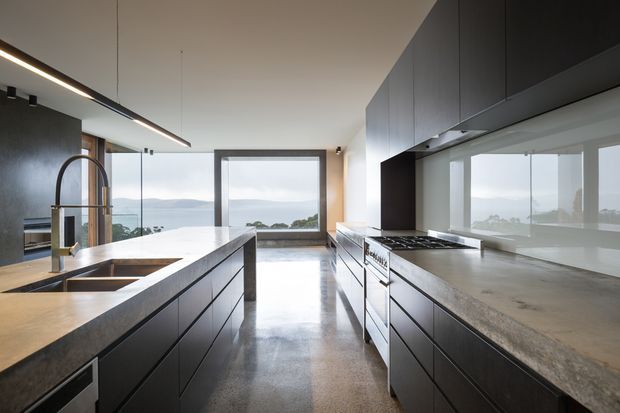 A spacious contemporary kitchen with a large central island, integrated double sink and sculptural tap, dark flat-panel cabinetry, a broad stone benchtop, and a built-in gas cooktop along the right wall. Full-height glazing and a large window provide expansive water views, while polished concrete flooring and linear pendant lighting reinforce the minimalist modern character.