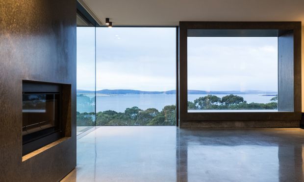 A contemporary interior living space with expansive floor-to-ceiling glazing overlooking a calm coastal bay and distant hills. The room features a polished concrete floor, dark built-in wall surfaces, a recessed fireplace, and a large dark-framed picture window, creating a minimalist, dramatic connection to the landscape.